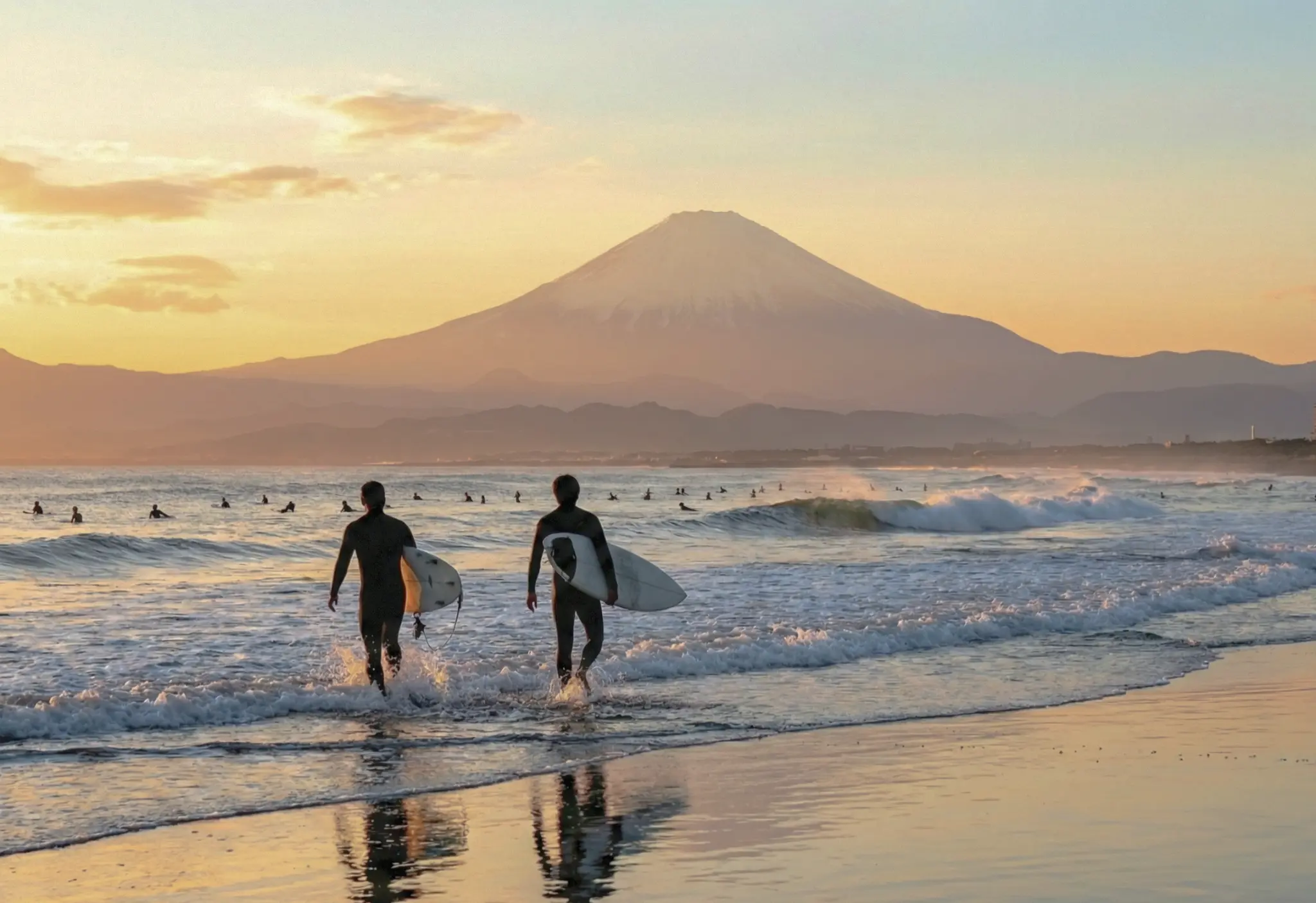 Two surfers carrying their boards walk into the ocean at sunset, with Mount Fuji rising behind the lineup near Tokyo