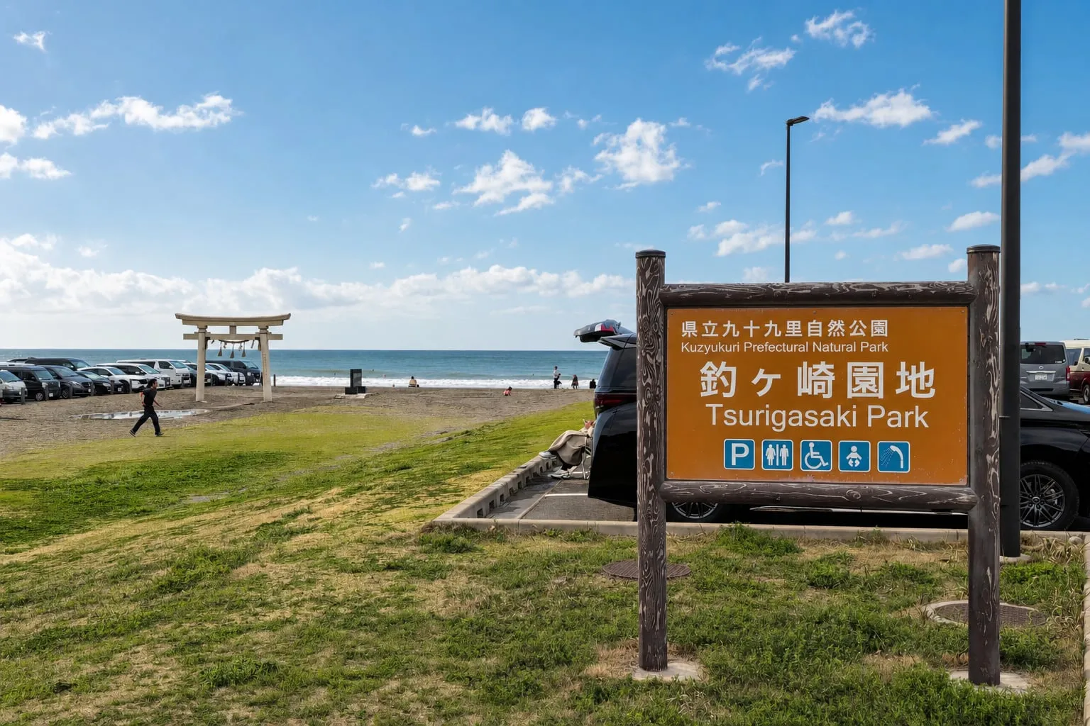 The Tsurigasaki Park sign at the Tokyo 2020 Olympic surfing venue in Chiba, with a white torii gate, beach parking, and the Pacific Ocean on a sunny day.